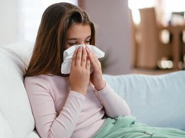 Young female sitting on the couch, blanket over her legs and blowing her nose.